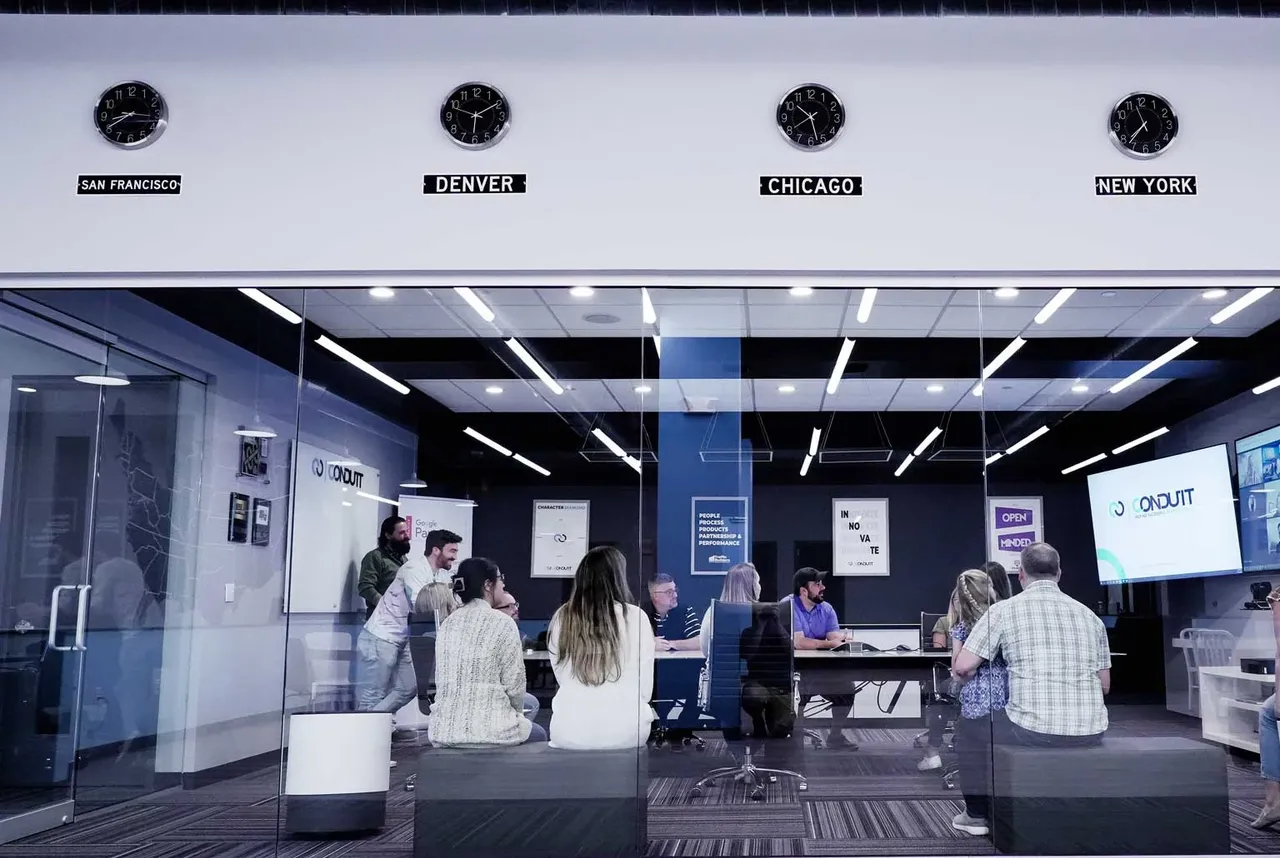 The Conduit conference room behind glass, four time-zone clocks above: San Francisco, Denver, Chicago, New York