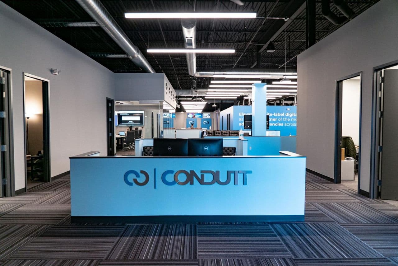 The Conduit reception desk carrying the company logo, looking through to the open office floor and the brand wall beyond