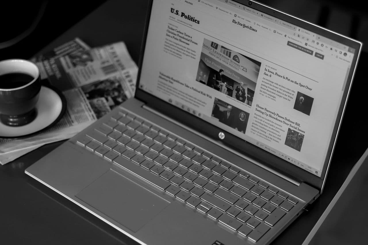 Black and white image of a laptop displaying news articles, accompanied by a cup of coffee and newspapers
