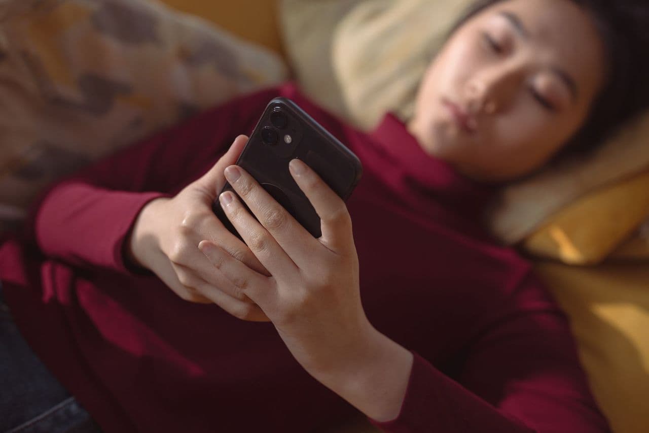 A young woman lying down, holding and using a smartphone for relaxation indoors