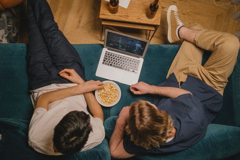 Two friends enjoying a movie on a laptop with popcorn from a cozy couch perspective