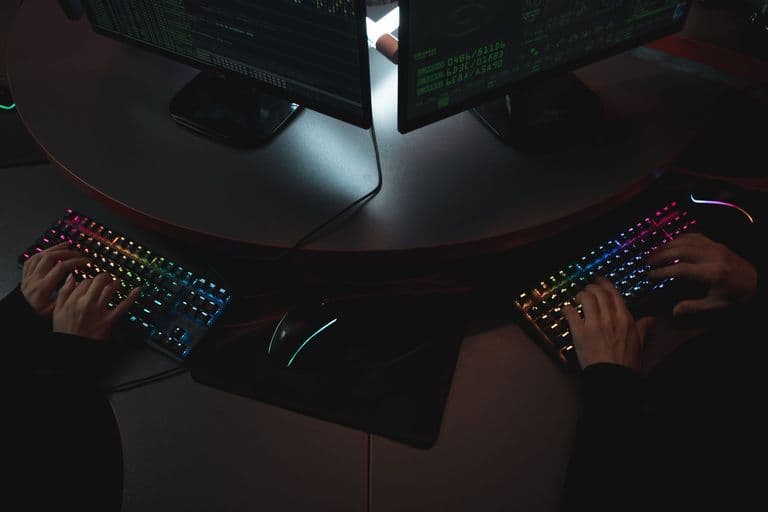 Two people typing on RGB keyboards with code on screens, indicating a cybersecurity environment