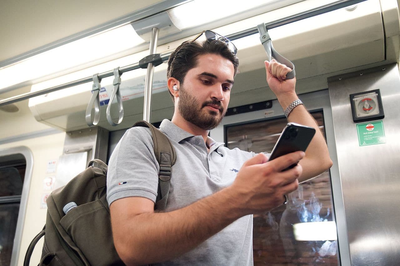A young man with a backpack uses his smartphone while standing on public transport, likely a metro or tram